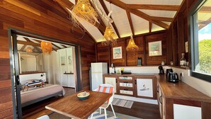 Ein gemütliches Blockhaus-Interieur mit Holzbalkendecke, einer Essecke mit Tisch und Stühlen, einer Küche mit Kühlschrank und Kaffeemaschine und einem großen Fenster mit Blick ins Freie.