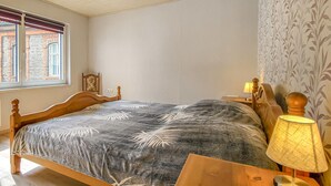 Une chambre avec un lit en bois, une table de chevet avec une lampe, une fenêtre avec des stores et un papier peint à motifs sur le mur.