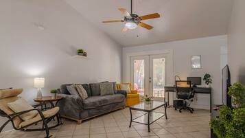 Ein Wohnzimmer mit einer Couch, einem Schaukelstuhl, einem Couchtisch, einem Schreibtisch mit Computer und einem Fernseher.