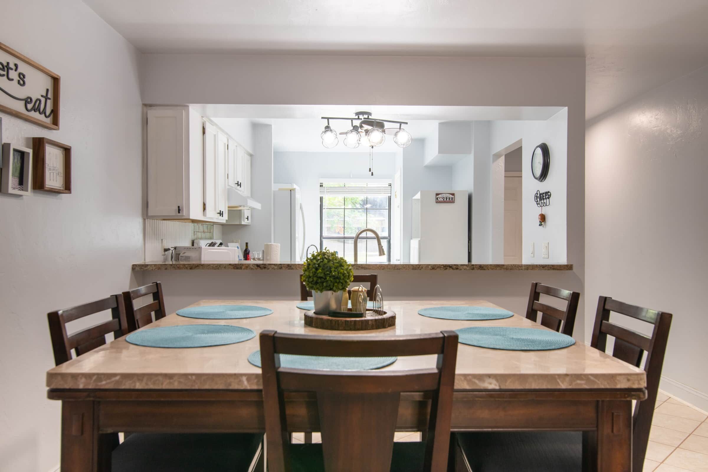 A dining room with a large mirror, wooden chairs, a central table with a plant, and a kitchen visible through the mirror.