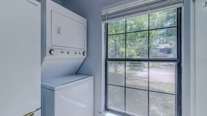 A laundry room with a front-loading washer and dryer, a window with blinds, and a view of a residential area outside. - Gainesville Cozy Pet friendly Near UF & Downtown (Gainesville)