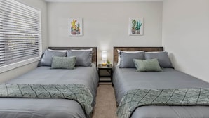 A bedroom with two beds, a nightstand, a lamp, and two wall-mounted cactus paintings. - Gainesville Nook Room (Gainesville)