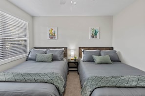 A bedroom with two beds, a nightstand, a lamp, and two wall-mounted cactus paintings. - Gainesville Nook Room (Gainesville)