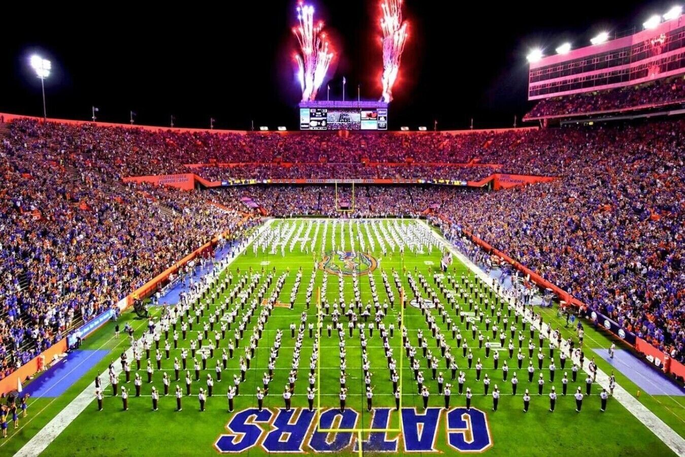 A football stadium with a marching band formation on the field, surrounded by a large crowd.