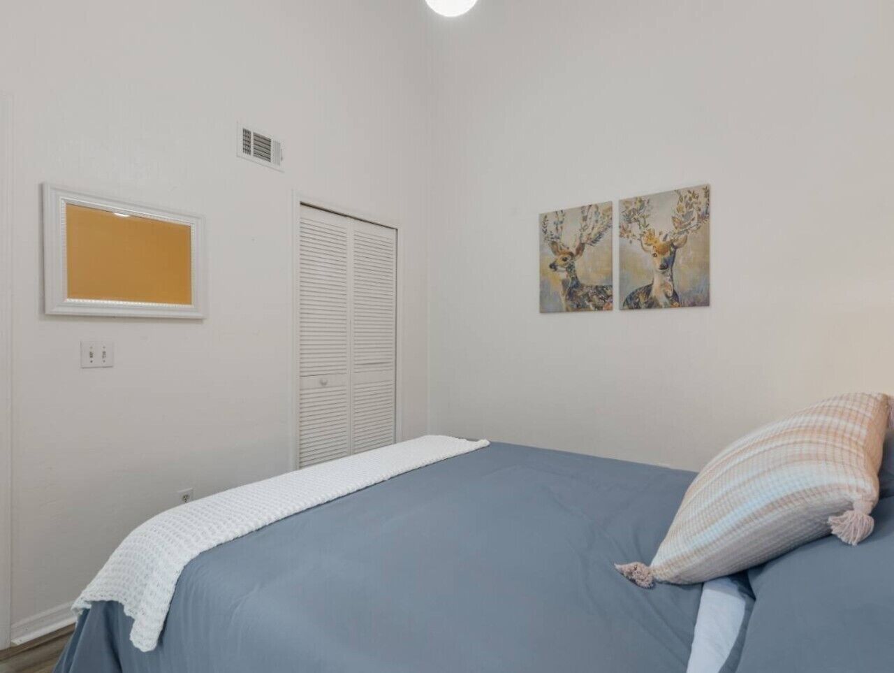 A bedroom with a bed, a white closet, a window with blinds, and two wall-mounted paintings.
