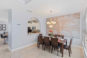 Uma área de refeições com uma mesa posta para quatro pessoas, um candelabro e uma cozinha visível através de um arco.