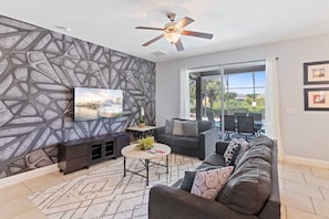 Uma sala de estar moderna com um design geométrico na parede, um ventilador de teto, um sofá, uma mesa de centro e uma televisão.