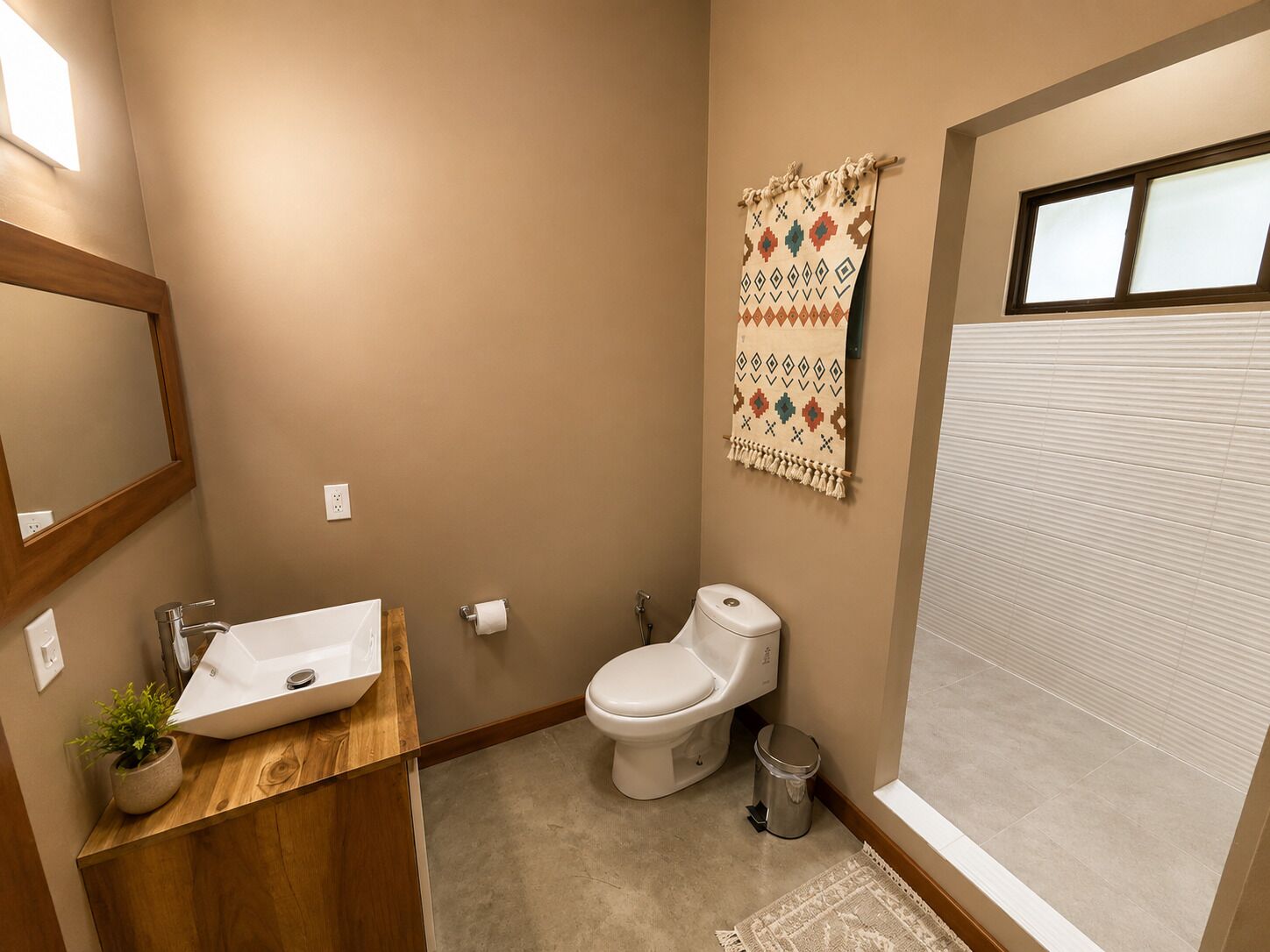 A bathroom with a wooden vanity, a white sink, a toilet, a trash can, and a patterned towel on the wall.