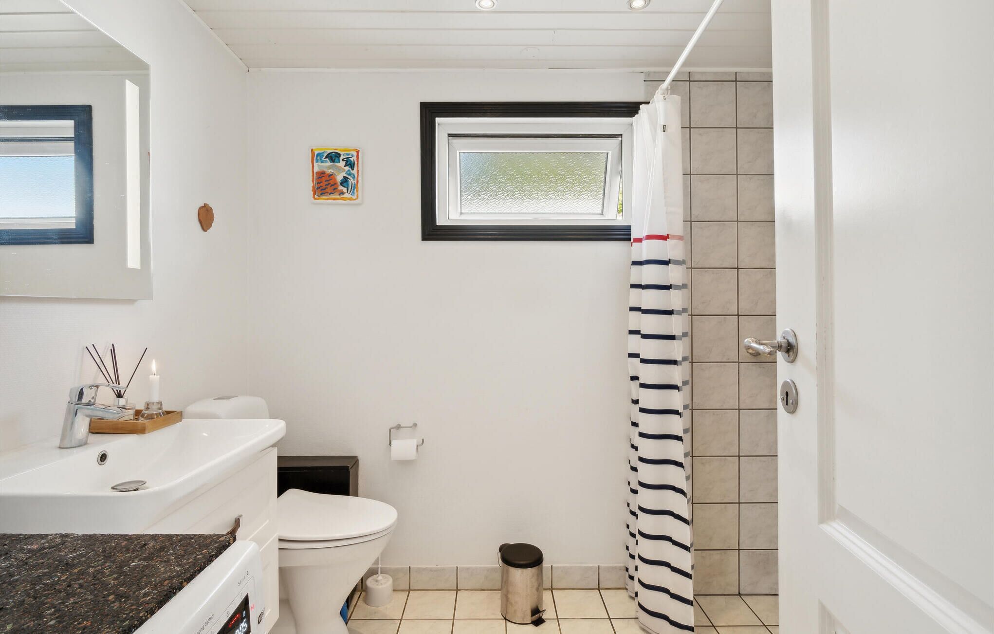 Une salle de bain avec des toilettes, un lavabo et une douche. Il y a une fenêtre au-dessus des toilettes et un petit tableau accroché au mur.