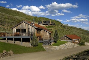 A rustic wooden cabin with a porch, a smaller adjacent building, and a mountainous backdrop.