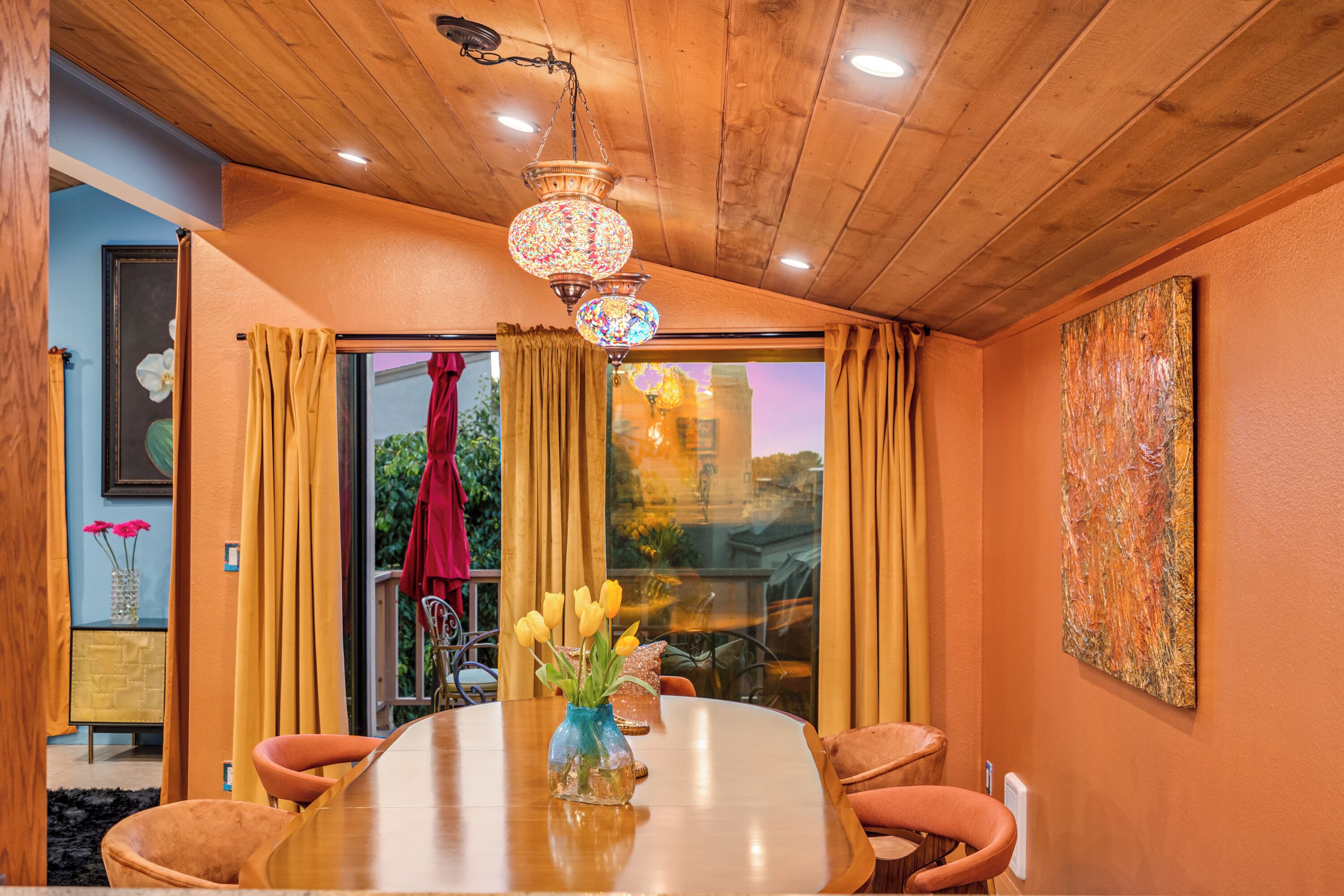 A dining area with a wooden table, chairs, a vase with flowers, a chandelier, and a view through a glass door.