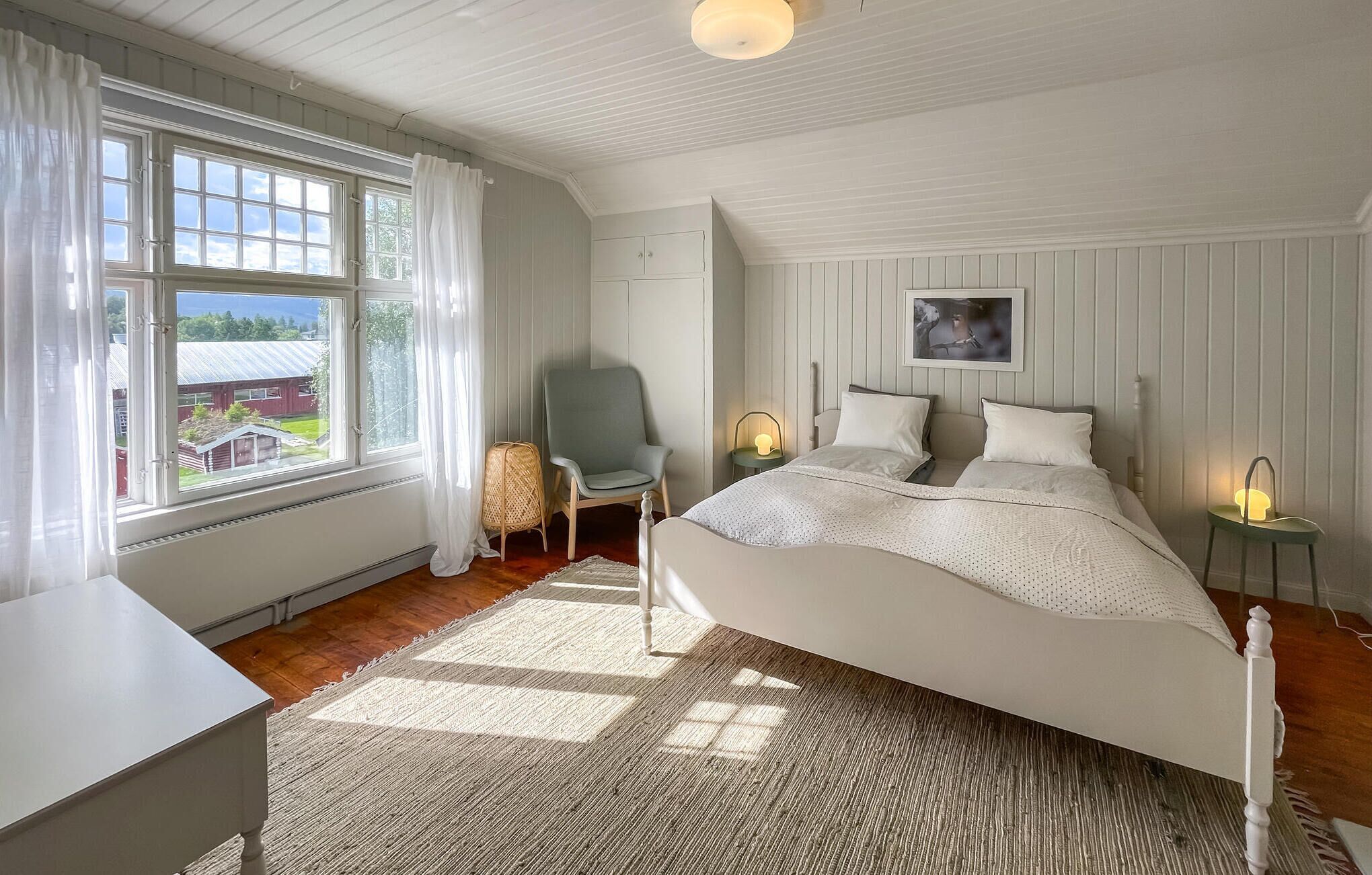 A bedroom with a large bed, a chair, a desk, and a window with curtains.