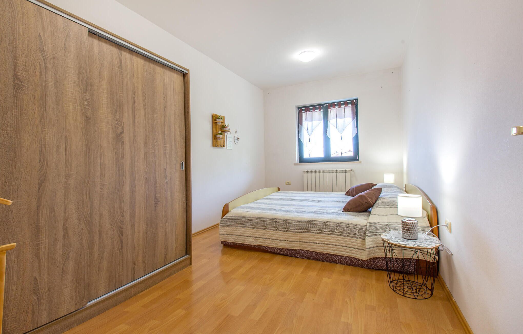 A bedroom with a large wooden wardrobe, a bed with a striped duvet, a bedside table with a lamp, and a window with curtains.