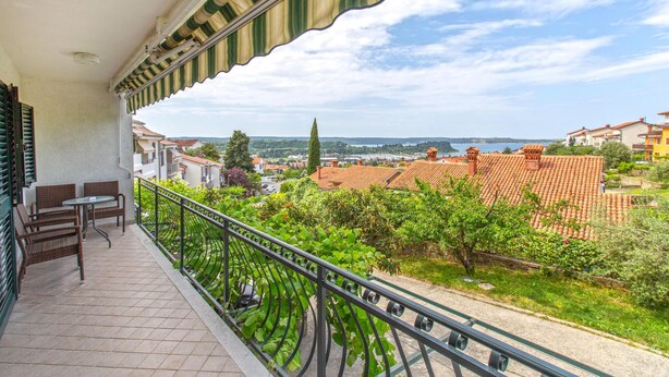 A balcony with wicker furniture, a metal railing, and a view of a town with red-roofed buildings and greenery.