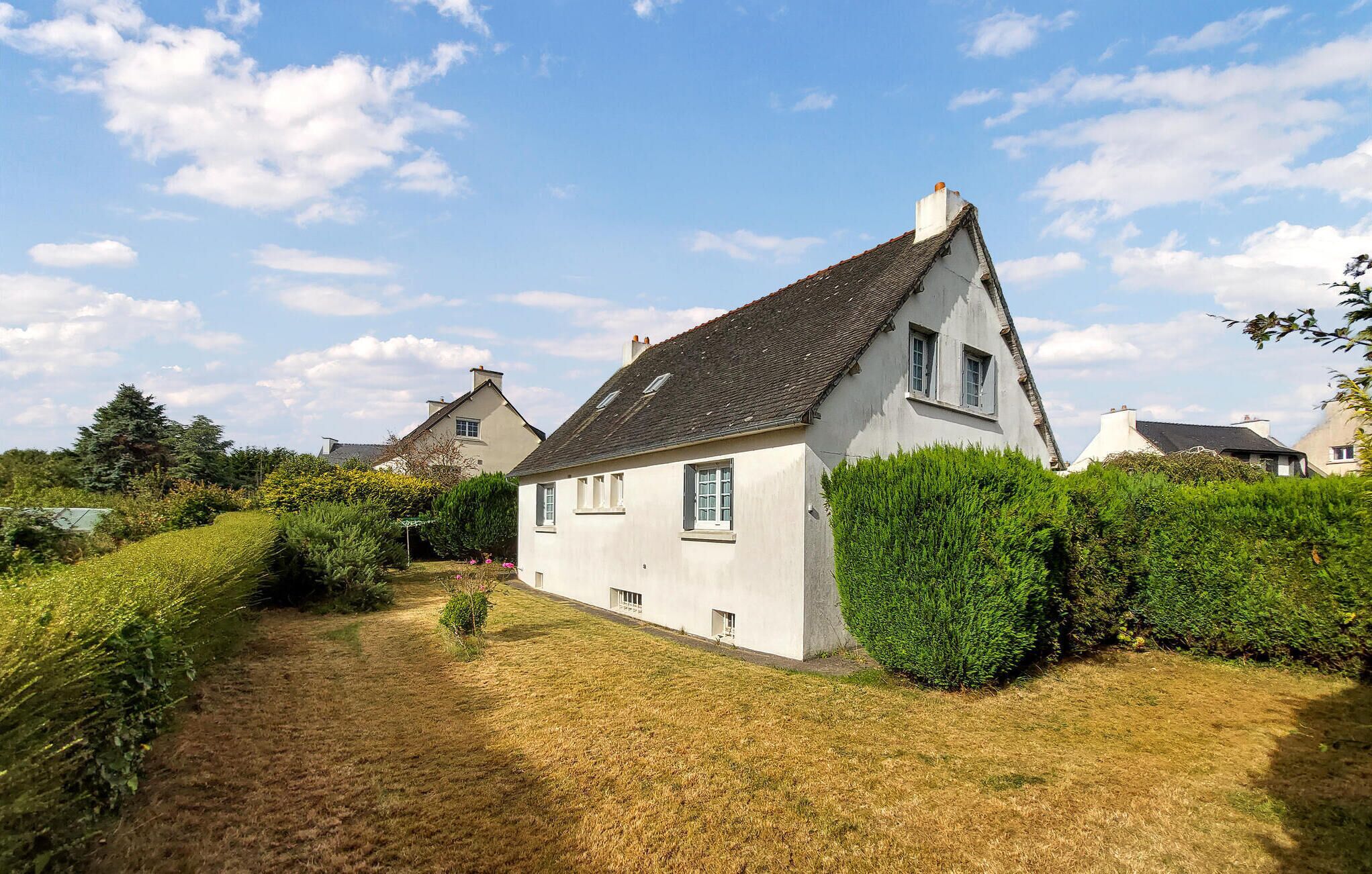 Une maison blanche avec un toit à forte pente, entourée de haies bien entretenues et d’une pelouse.