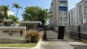 Un moderno edificio de gran altura con entrada protegida por un portón, rodeado de vegetación y palmeras.