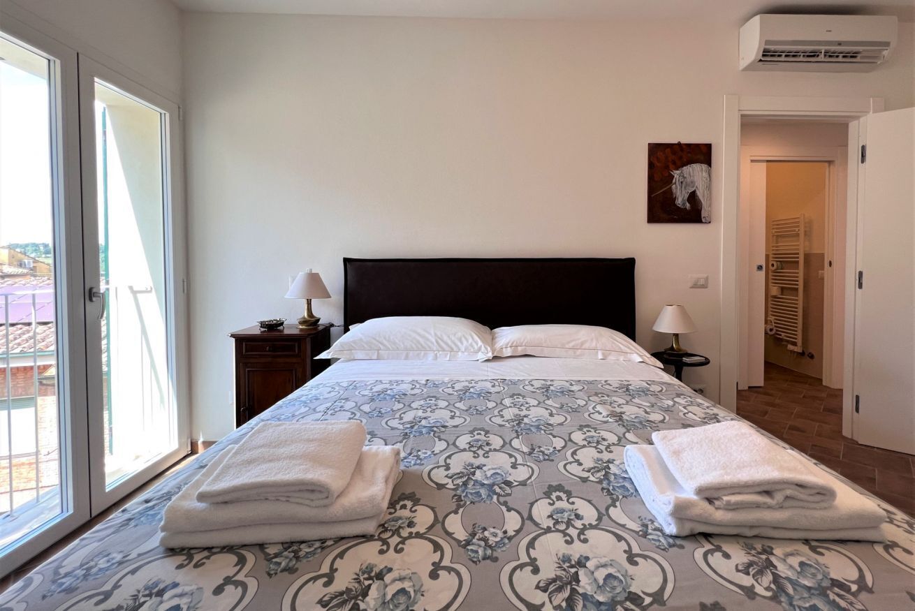 Una cama bien hecha con una colcha estampada, dos almohadas blancas y toallas dobladas sobre la cama. Hay una mesita de noche con una lámpara y una pequeña mesa con otra lámpara. Un cuadro cuelga en la pared sobre la cama.