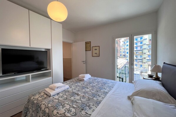 A hotel room with a large bed, a flat-screen TV, a balcony with a view of buildings, and a door leading to a bathroom.