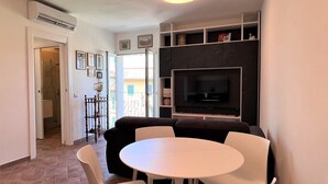 Un salon moderne avec un canapé noir, des fauteuils blancs, une table ronde et un téléviseur fixé à un mur foncé.