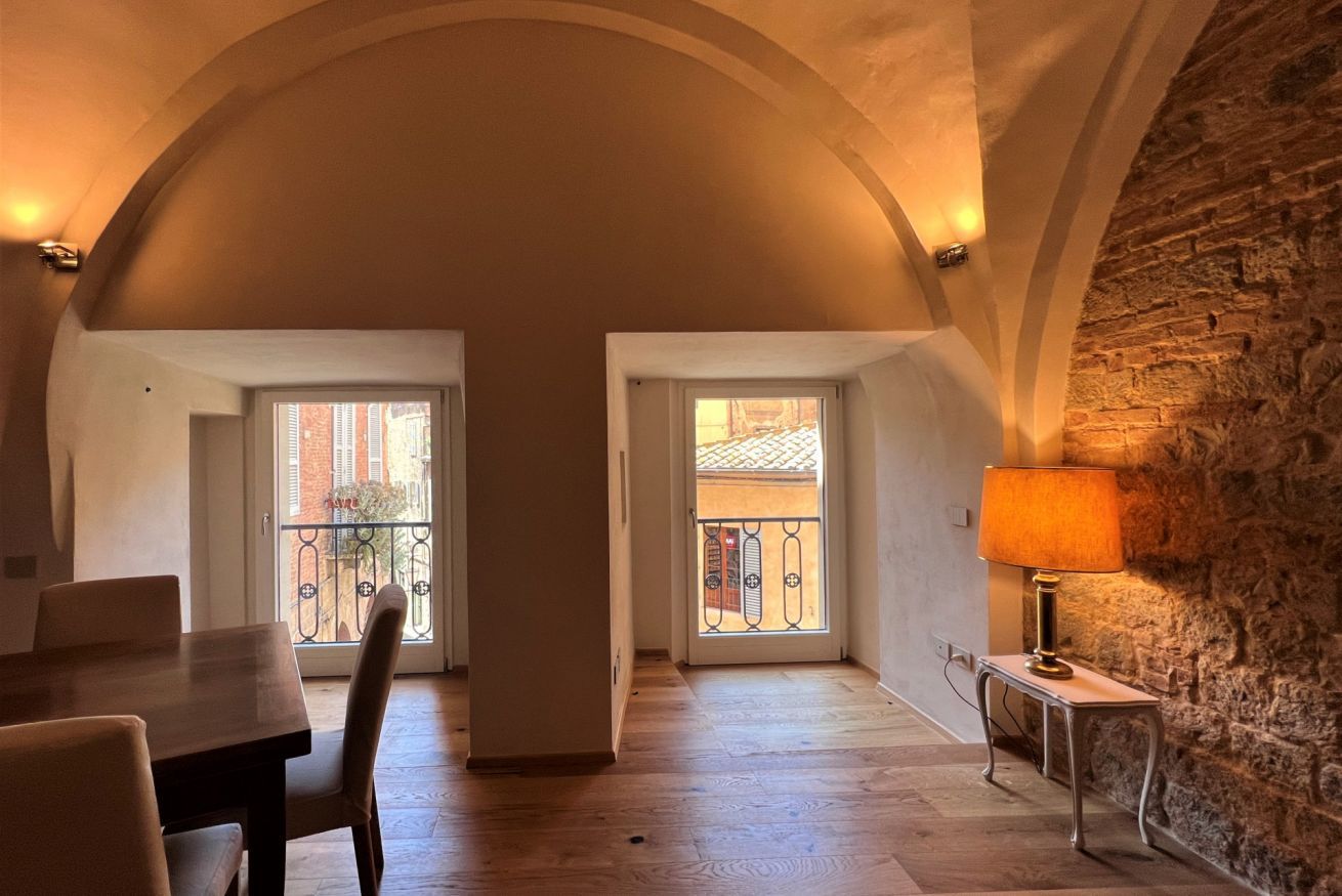 A spacious room with a wooden dining table and chairs, a stone wall, and a view through a window to a balcony.