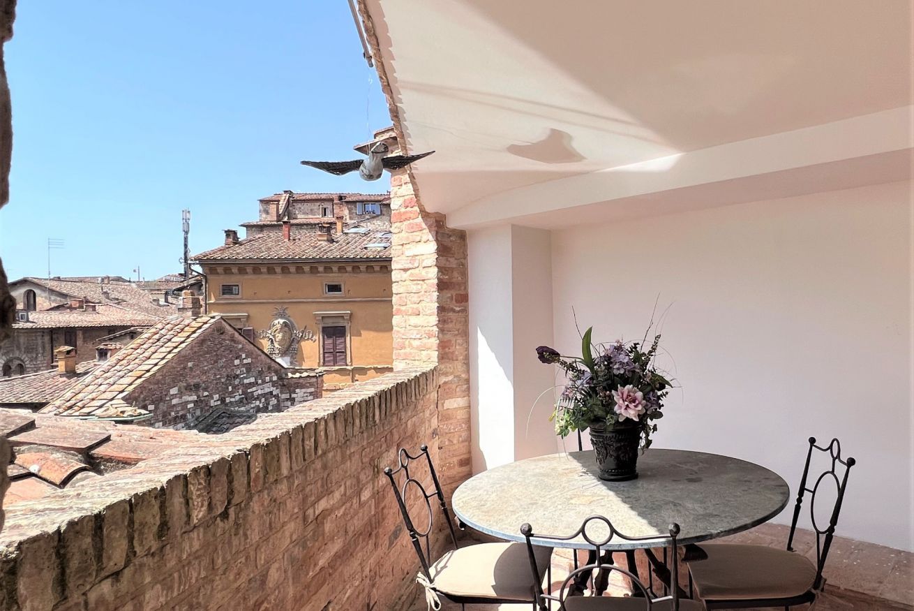 Mesa redonda con cuatro sillas, una planta en maceta y una escultura de pájaro en un balcón con vistas a un paisaje urbano con edificios históricos.