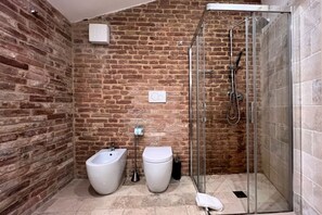 A bathroom with a brick wall, a glass-enclosed shower, a bidet, and a toilet. - Tolomei Terrace (Siena)