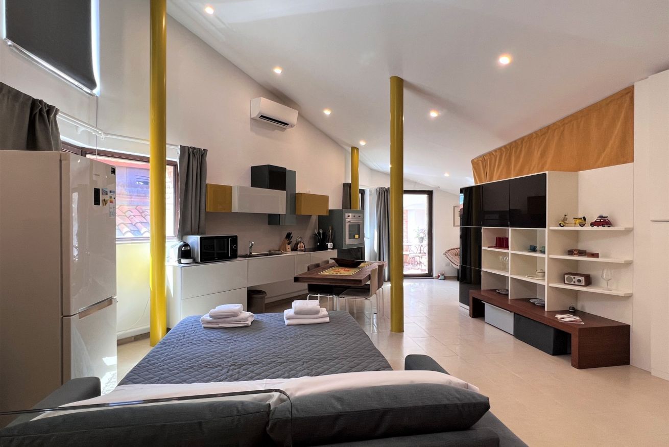 A modern hotel room with a bed, a desk, a kitchenette, and a TV.