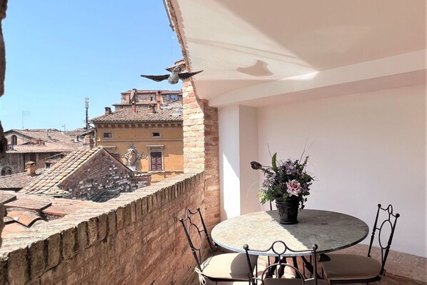 A round table with four chairs, a potted plant, and a bird sculpture on a balcony overlooking a cityscape with historic buildings. - Tolomei Terrace (Siena)