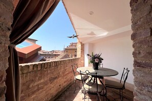 A balcony with a round table and chairs, a potted plant, and a view of buildings in the background. - Tolomei Terrace (Siena)