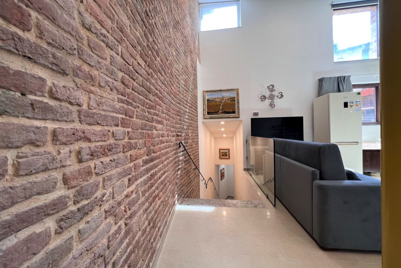 A staircase with a brick wall on one side and a modern living area with a sofa and a flat-screen TV on the other.