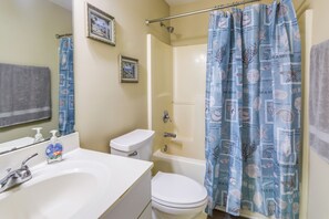 Un baño con lavamanos, inodoro y bañera blancos. Hay una cortina de ducha con estampado de conchas marinas.