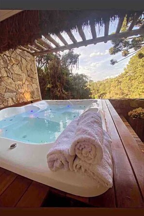 A hot tub with a stone wall and wooden deck, surrounded by greenery and a clear sky.