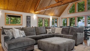 Ein geräumiges Wohnzimmer mit einer großen Eckcouch, mehreren Fenstern mit Blick auf Bäume und einer Holzbalkendecke.