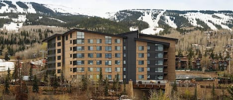 Un hotel de varios pisos con diseño moderno, que combina madera y paneles oscuros, con vistas a montañas nevadas y un cielo despejado.