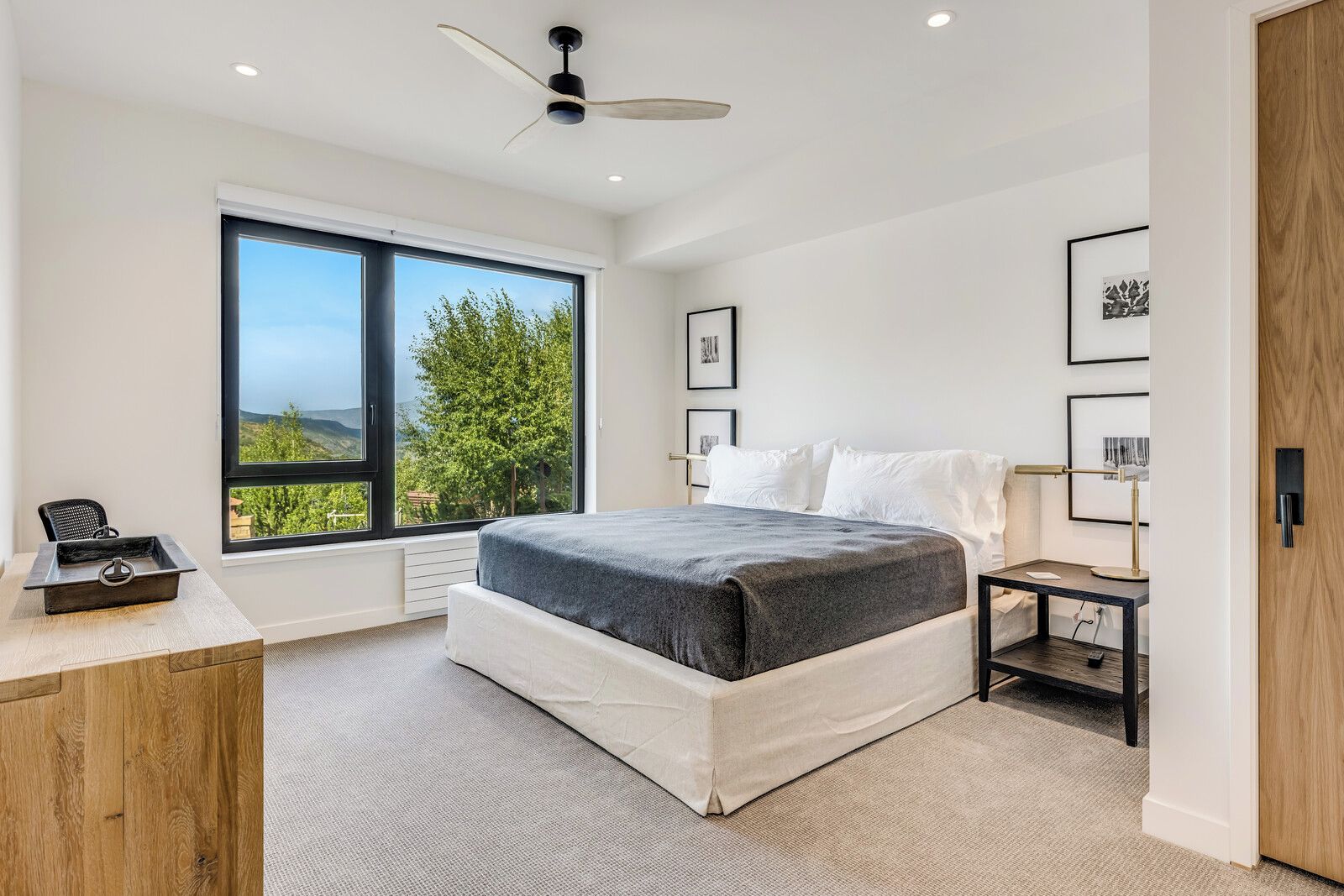 A bedroom with a large window, a bed, a desk, and a nightstand.