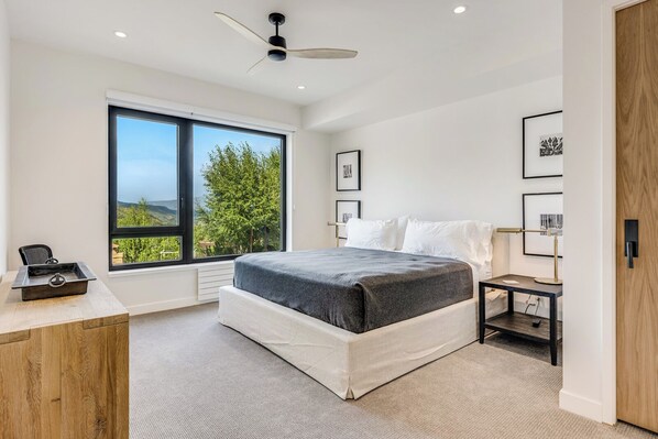 A bedroom with a large window, a bed, a desk, and a nightstand.