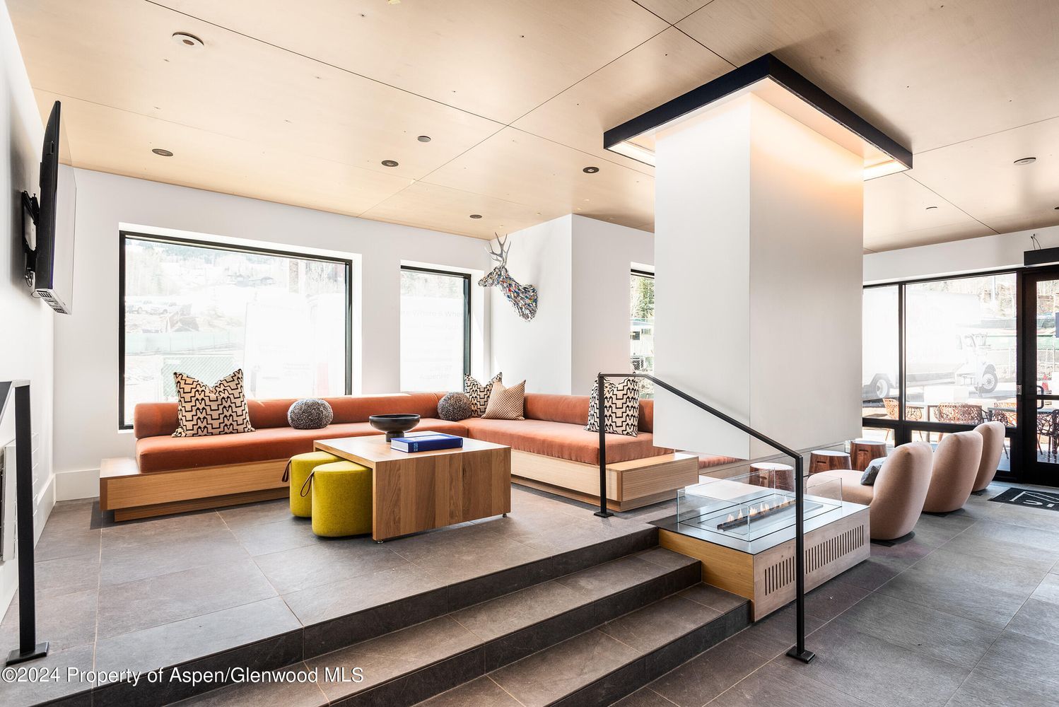 Ein modernes Wohnzimmer mit Sofa, Hocker und Couchtisch, einer Treppe und großen Fenstern.
