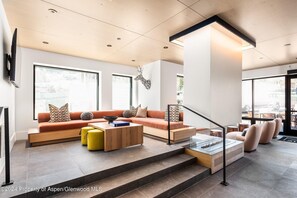 Ein modernes Wohnzimmer mit Sofa, Hocker und Couchtisch, einer Treppe und großen Fenstern.
