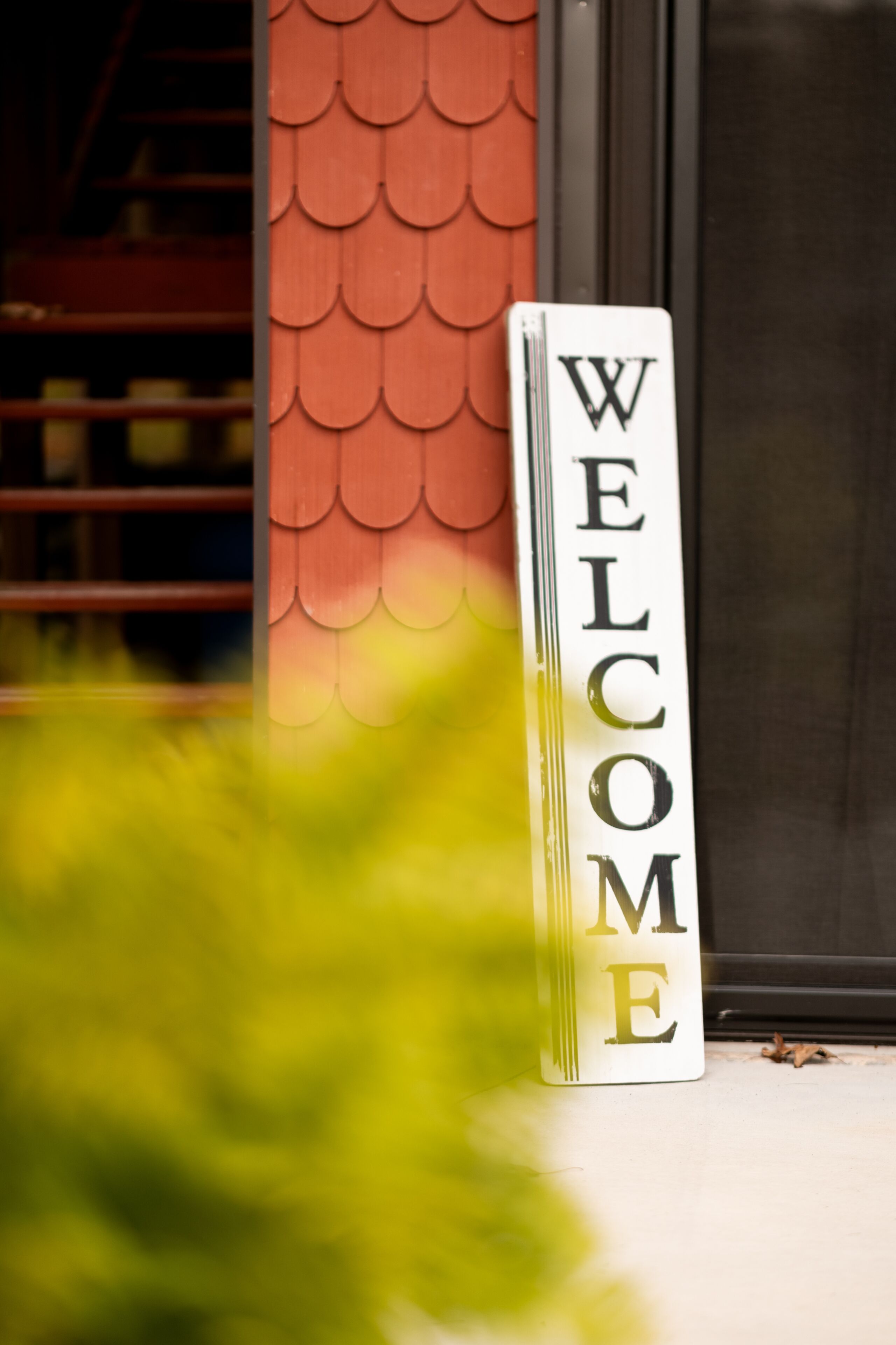 A vertical welcome sign with the word "WELCOME" in large letters, placed on a white background, next to a red textured wall and a glass door.