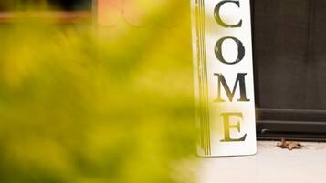 A vertical welcome sign with the word "WELCOME" in large letters, placed on a white background, next to a red textured wall and a glass door.