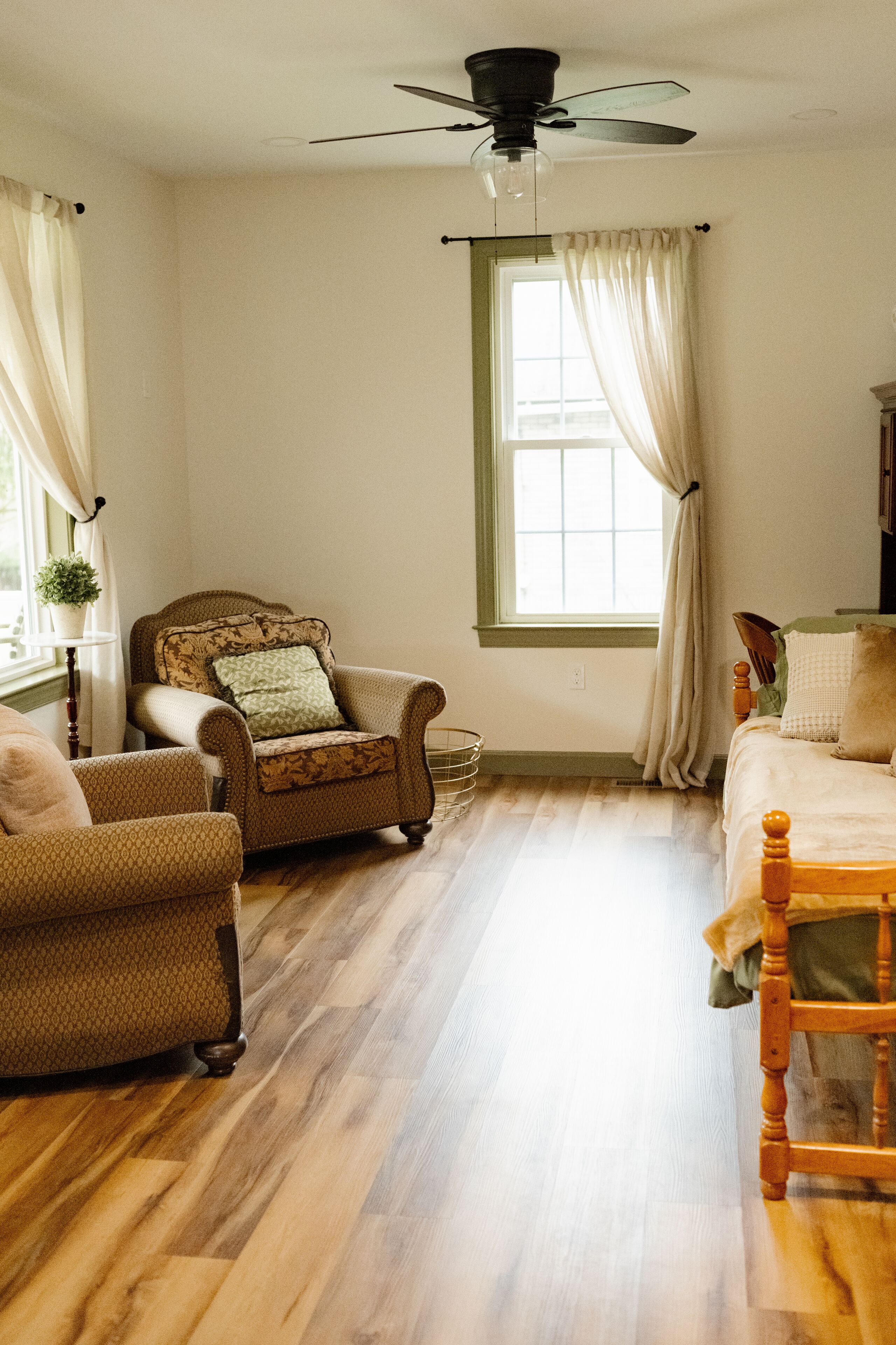 A room with a wooden floor, a ceiling fan, a window with curtains, a bed, and two armchairs.