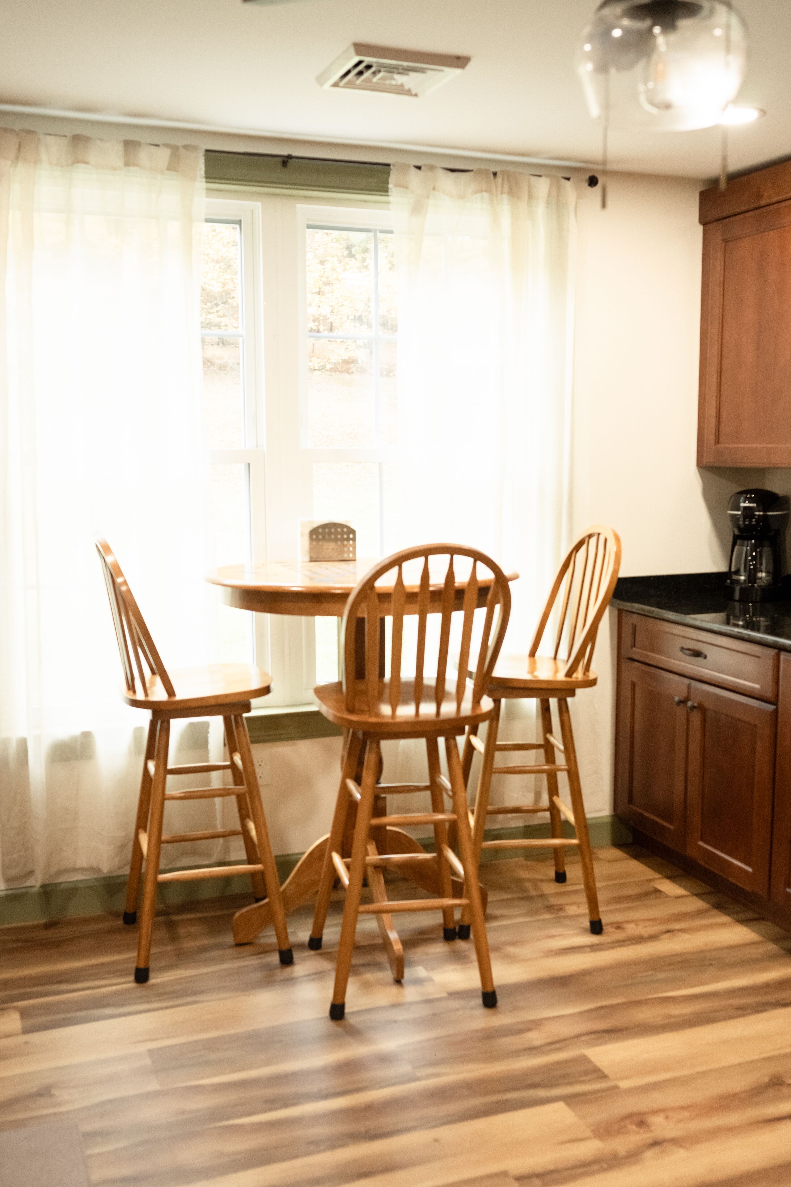 A dining area with a round table and four wooden chairs, a window with curtains, and wooden cabinets.