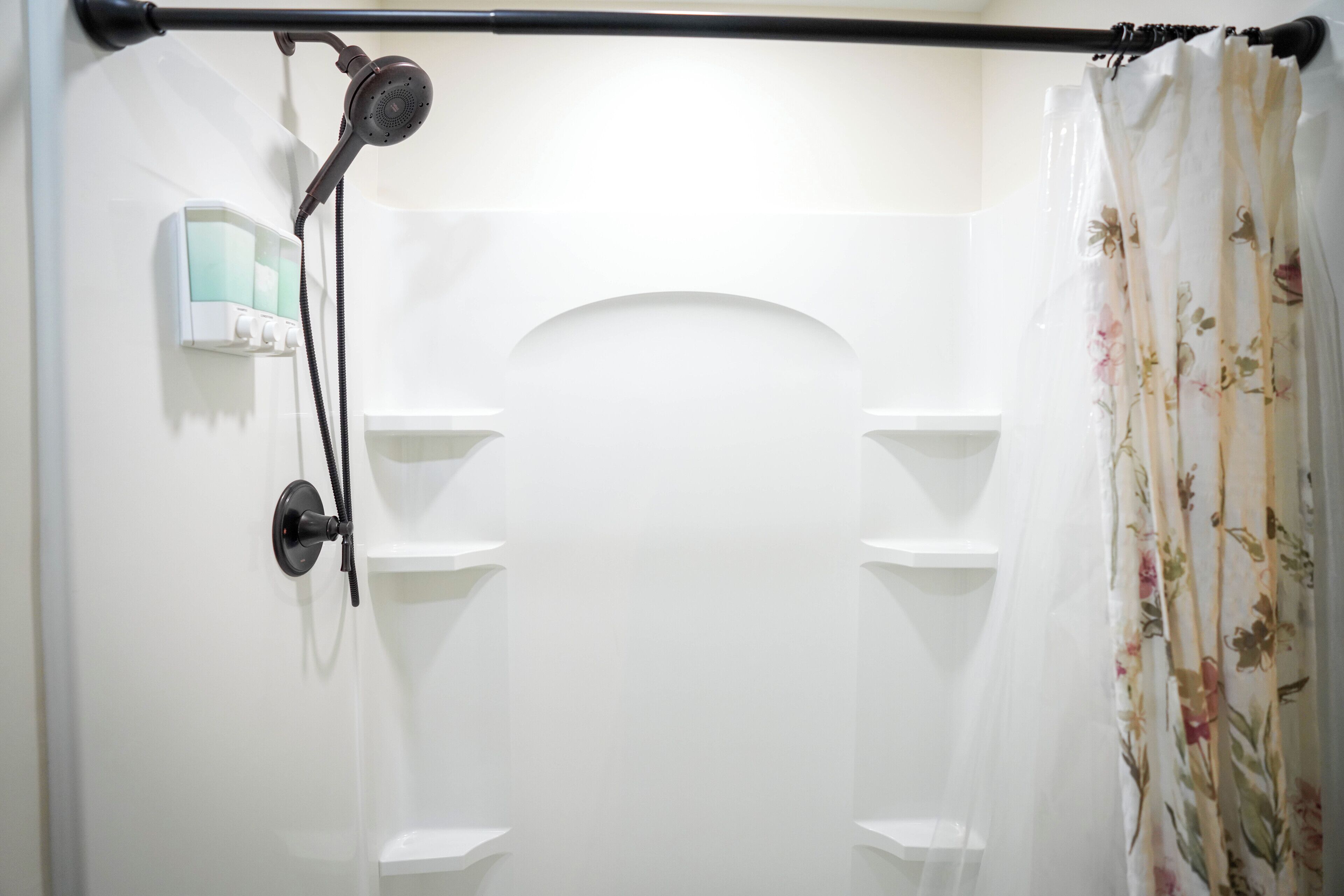 A shower area with a handheld showerhead, a soap dispenser, and a floral shower curtain.
