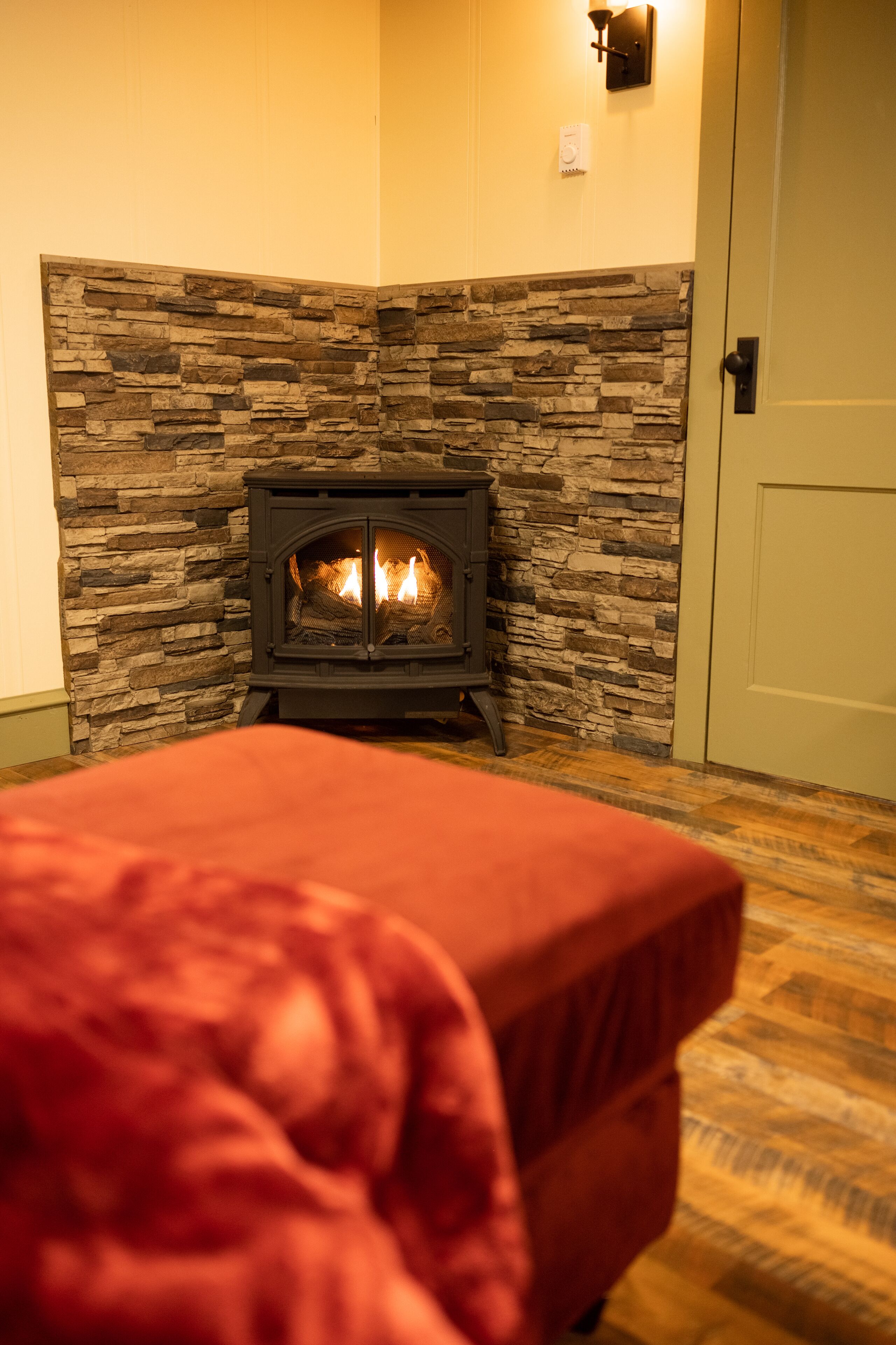 Una habitación con chimenea de piedra, una cama con manta roja y una puerta a la derecha.
