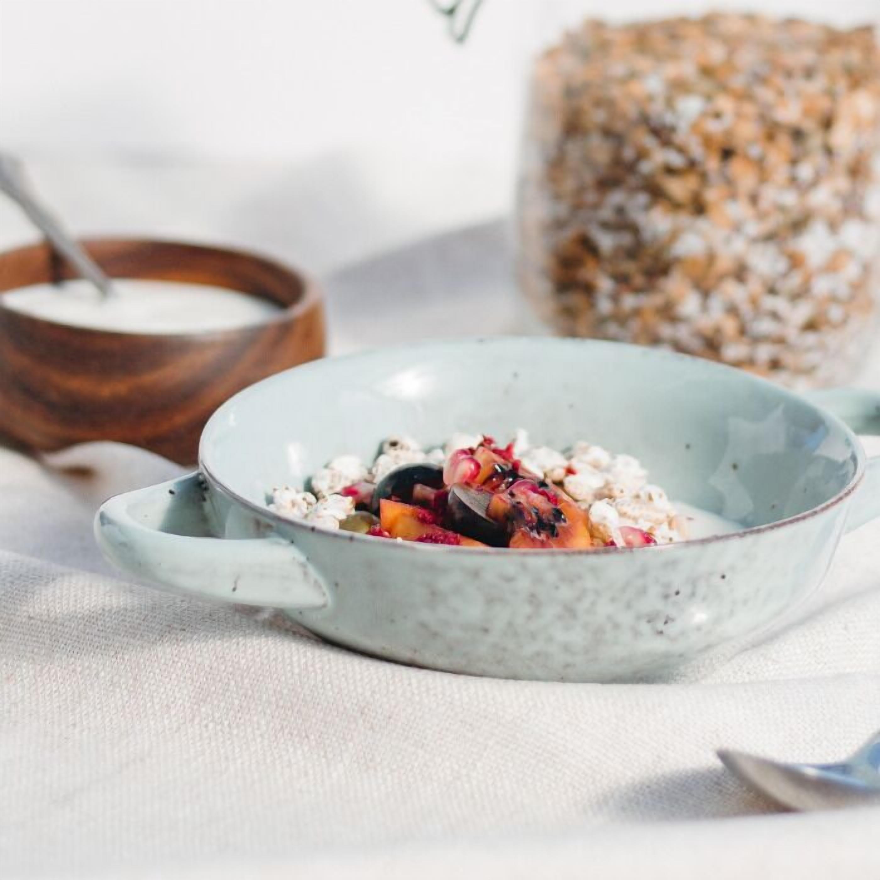 A bowl of cereal with fruits and a wooden bowl of milk in the background.