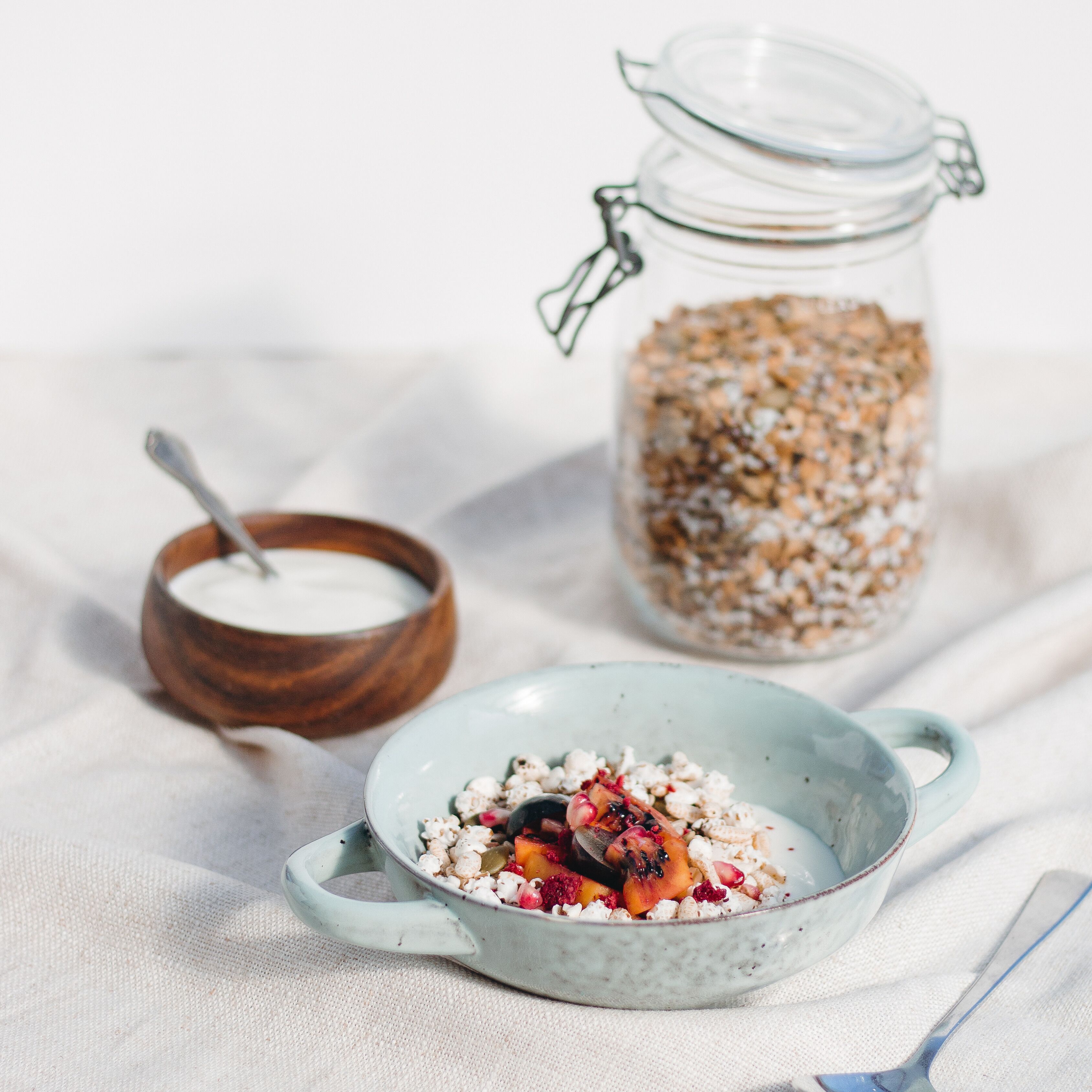 A bowl of cereal with fruits and nuts, a jar of granola, and a wooden bowl with a spoon.