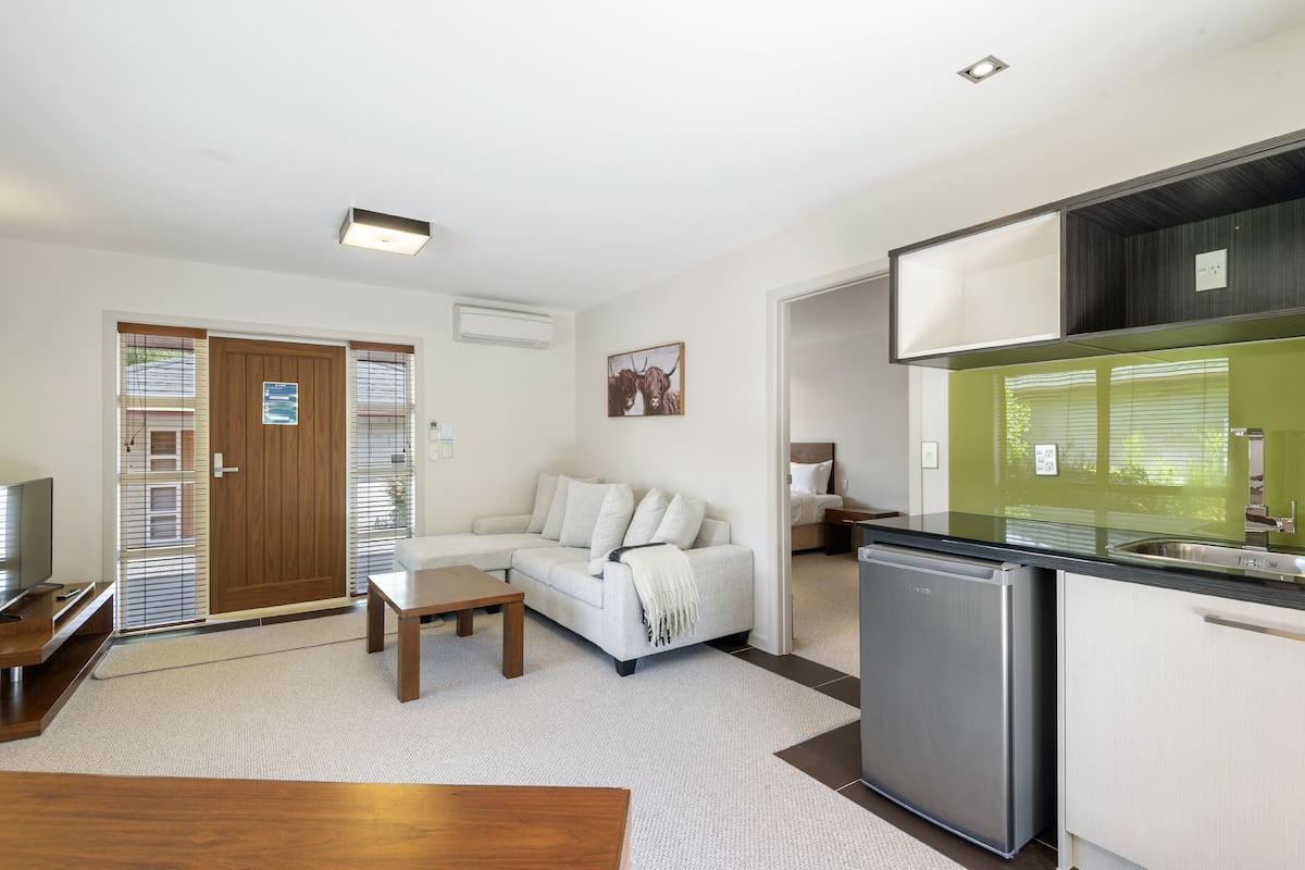 Una sala de estar moderna con un sofá, una mesa de centro y un televisor. Hay una zona de cocina con un refrigerador y un lavamanos.