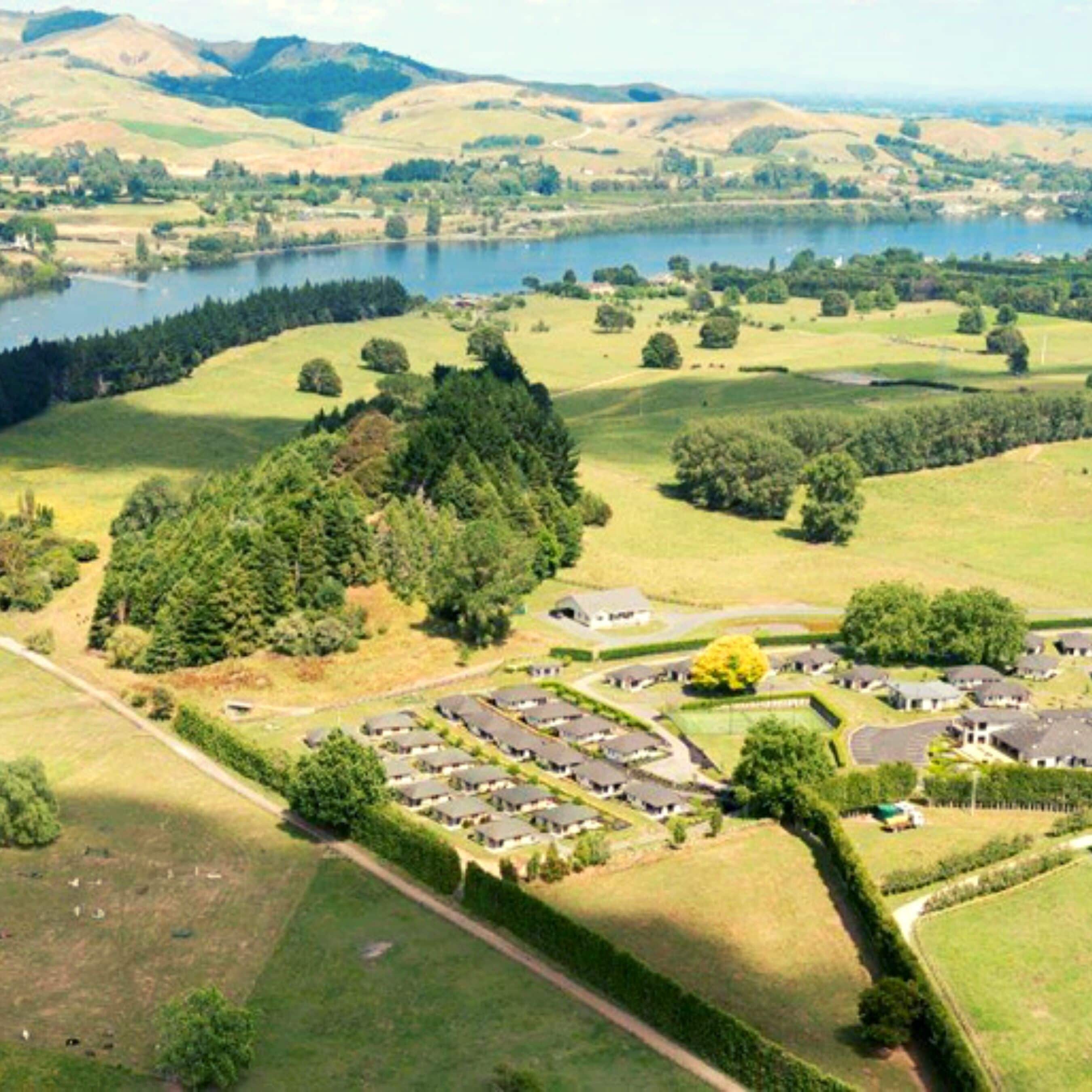 A residential area with houses arranged in a semi-circular pattern around a central green space, with a lake and hills in the background.