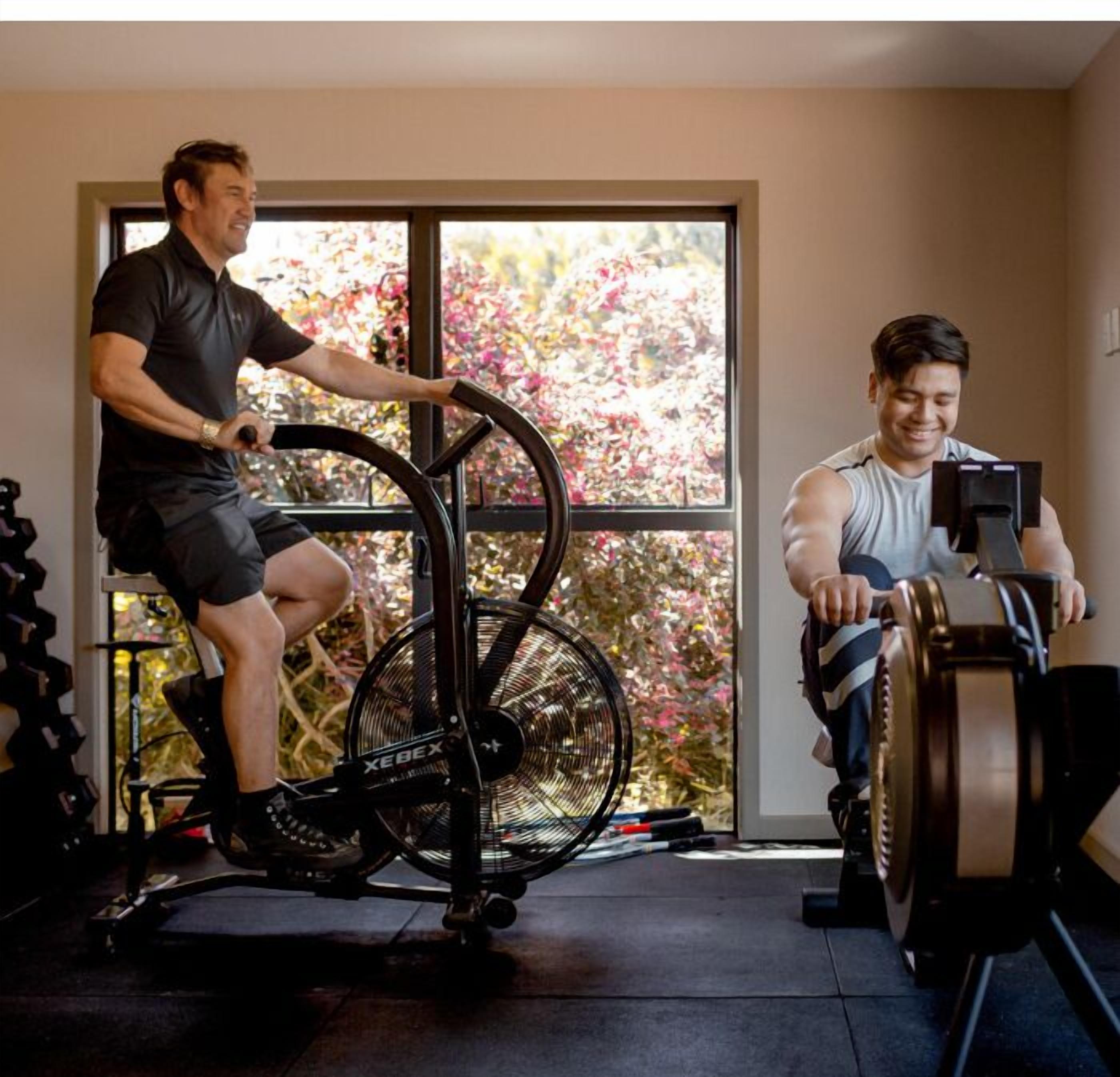 Two people using stationary bikes in a room with large windows and a view of trees.