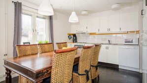Un comedor con mesa de madera y sillas de mimbre, una cocina con armarios blancos y un ventanal con cortinas.
